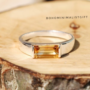 Puede incluir: Un anillo de plata con una piedra rectangular de color naranja claro engastada en una banda de plata. El anillo está sobre una superficie de madera.