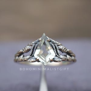 May include: A silver ring with a clear, kite-shaped gemstone. The ring has a detailed, leafy design around the gemstone and band. The ring is set against a blurred background.