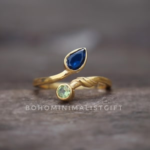 May include: A gold-colored ring with a leaf-like design. The ring features a teardrop-shaped, dark blue gemstone and a round, light green gemstone. The ring's band has a twisted rope design. The text "BOHOMINIMALISTGIFT" is visible.