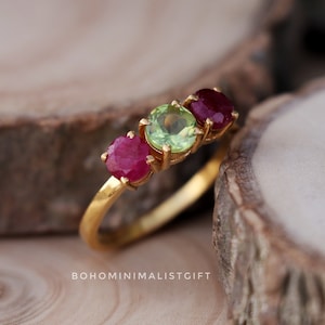 Puede incluir: Un anillo de oro con tres piedras preciosas: dos rubíes rojos y un peridoto verde. El anillo está sobre una superficie de madera.