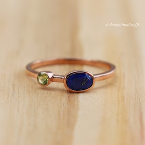 Może przedstawiać: Złoty pierścionek z niebieskim owalnym kamieniem lapis lazuli i mniejszym zielonym owalnym kamieniem perydot.