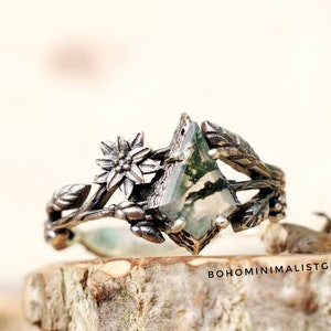 May include: A silver-toned ring featuring a geometric, green moss agate stone. The ring is adorned with floral and leaf details, creating a nature-inspired design. The ring is set on a wooden surface. The text "BOHOMINIMALISTGIFT" is visible.