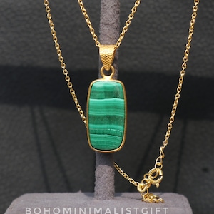 Puede incluir: Un collar dorado con un colgante rectangular con una piedra de malaquita verde vibrante. La piedra está engastada en un marco dorado y cuelga de una fina cadena. El collar se exhibe sobre un soporte gris. El texto "BOHOMINIMALISTGIFT" es visible.