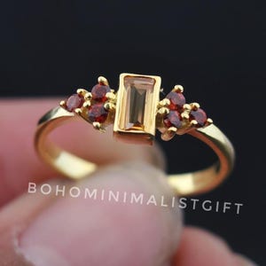 Może przedstawiać: Złoty pierścionek z prostokątnym, jasnobrązowym kamieniem szlachetnym pośrodku, otoczonym grupami mniejszych, czerwonych kamieni. Pierścionek ma delikatny pierścień. Widoczny jest napis "BOHOMINIMALISTGIFT".