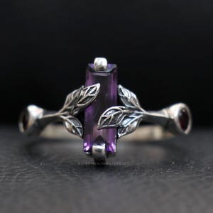 Könnte beinhalten: Ein silberner Ring mit einem rechteckigen, tiefvioletten Edelstein als Mittelpunkt. Der Ring ist mit blattartigen Details auf beiden Seiten des Edelsteins und kleinen, runden roten Edelsteinen an den Enden verziert. Der Ring ist vor dunklem Hintergrund.