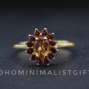 Peut inclure: Une bague en or ornée d'une pierre centrale ovale de couleur ambre, entourée de petites pierres rouge foncé. La bague présente un anneau fin et un motif floral. Le texte "BOHOMINIMALISTGIFT" est visible en bas.
