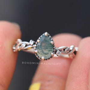 May include: A silver ring with a teardrop-shaped moss agate gemstone. The ring has a detailed vine and leaf pattern on the band, with small clear stones. The text "BOHOMINIMALISTGIFT" is visible.