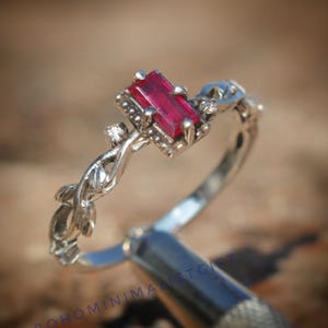 Könnte beinhalten: Ein silberner Ring mit einem rechteckigen, tiefroten Edelstein. Der Ring hat ein filigranes, verziertes Design mit kleinen Akzentsteinen und einem weinrebenartigen Muster am Band. Der Ring wird vor einem unscharfen, natürlichen Hintergrund gezeigt.