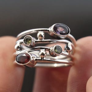 May include: A multi-band sterling silver ring featuring various oval and round gemstones in shades of purple, green, and red. The ring has a unique design with small silver spheres between the gemstones.
