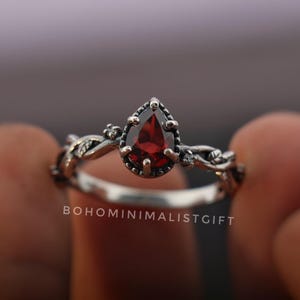 May include: A silver ring with a teardrop-shaped, deep red gemstone. The ring features a detailed, vine-like design around the band and the gemstone setting. The text "BOHOMINIMALISTGIFT" is visible on the ring.