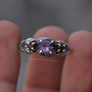 May include: A silver ring featuring three gemstones. The center stone is a round, light purple amethyst, flanked by two smaller, round, red gemstones. The ring has a floral design around the stones and the text "BOHOMINIMALISTGIFT".