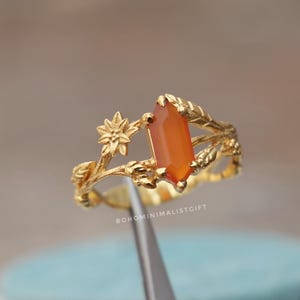 May include: A gold-colored ring with an orange gemstone. The ring features a floral and leaf design around the gemstone. The text "BOHOMINIMALISTGIFT" is visible on the ring.