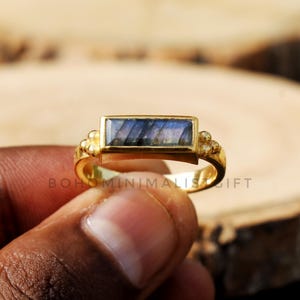 Pode incluir: Anel de ouro com uma pedra retangular de labradorita. O anel tem pequenas contas douradas nas laterais.