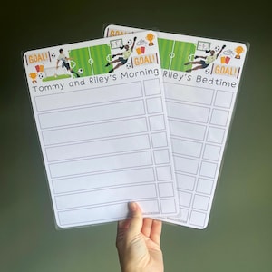 May include: Two laminated white charts with football-themed headers. One reads Tommy and Riley's Morning, the other Riley's Bedtime. Both feature illustrations of football players, goals, and trophies, with blank rows and checkboxes for daily tasks.
