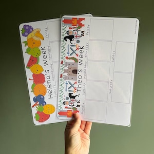 May include: Two rectangular laminated weekly planners with white backgrounds and black text. One features colourful cartoon fruit illustrations and the text Helena's Week. The other displays medieval knight and castle graphics with the text Theo's Week.
