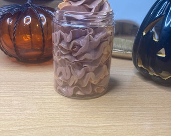 May include: A clear glass jar filled with a swirl of brown and tan whipped body butter. The jar is surrounded by Halloween decorations, including an orange glass pumpkin and a black pumpkin with a carved face.