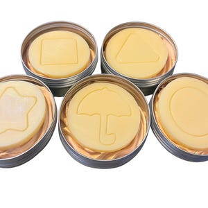 May include: Five light yellow, circular candies with embossed shapes, including a square, triangle, star, umbrella, and circle, are presented in individual silver tins. The candies rest on a light brown paper.