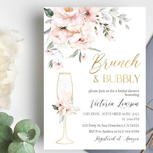 May include: A white invitation with gold lettering for a bridal shower. The invitation features a floral design with pink flowers and green leaves. The text reads "Brunch & Bubbly" and "Victoria Lowson".