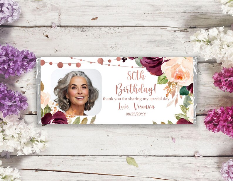 May include: A white chocolate bar wrapper with a floral design in shades of burgundy, pink, and green. The wrapper features a photo of a woman, the text "80th Birthday!" and "thank you for sharing my special day", and the signature "Love, Veronica" with the date "08/25/20YY".