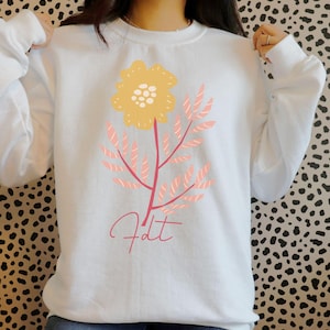 May include: White sweatshirt featuring a yellow flower with a light pink stem and leaves. The word "fdt" is written in a cursive font below the flower. The sweatshirt is a long-sleeved crew neck.