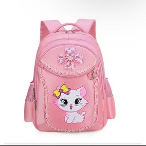 Pode incluir: Uma mochila rosa claro com um gato branco de desenho animado com um laço amarelo na cabeça. A bolsa inclui um laço decorativo rosa com lantejoulas no topo, acabamento com babados no bolso frontal, dois bolsos laterais de malha e alças ajustáveis.