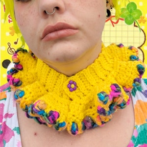 May include: A yellow crocheted collar with a colorful ruffled trim. The collar has a single yellow button with a purple flower design in the center.