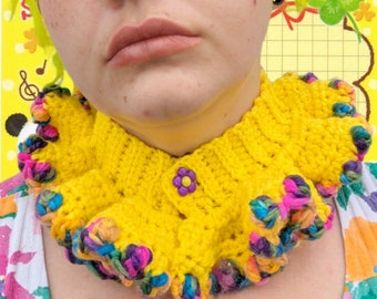 Può includere: Un colletto di uncinetto giallo con un bordo arricciato colorato. Il colletto ha un solo bottone giallo con un motivo a fiore viola al centro.