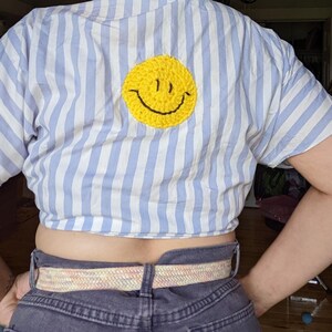May include: A blue and white striped shirt with a yellow smiley face embroidered on the back. The shirt is cropped and has a relaxed fit.