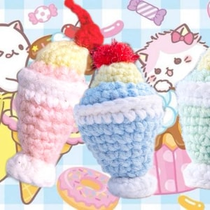 Puede incluir: Juguetes de sundae de helado de ganchillo en rosa, azul y verde con detalles blancos y adornos de cerezas rojas. El fondo presenta gatos de dibujos animados, plátanos, donas y dulces sobre un patrón de cuadros azules y blancos.