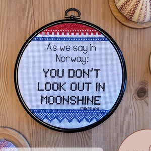 Puede incluir: Patrón de punto de cruz en formato PDF para un diseño que dice "Como se dice en Noruega: No mires a la luz de la luna". El diseño está en un aro de bordado negro con un borde rojo, blanco y azul. El diseño está sobre una superficie de madera con erizos de mar.