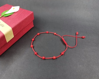 May include: Red beaded bracelet with adjustable cord closure. The bracelet features small, round red beads strung on a matching red cord. A red gift box with a white flower and the words "for you" is in the background.