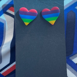 May include: A pair of heart-shaped stud earrings. Each earring features a rainbow design with horizontal stripes in red, orange, yellow, green, blue, and purple. The earrings are displayed on a black card.