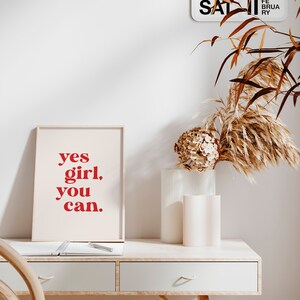 May include: A framed print with the text "yes girl, you can." in red letters on a white background. The print is sitting on a wooden desk with a white notebook and a pen.