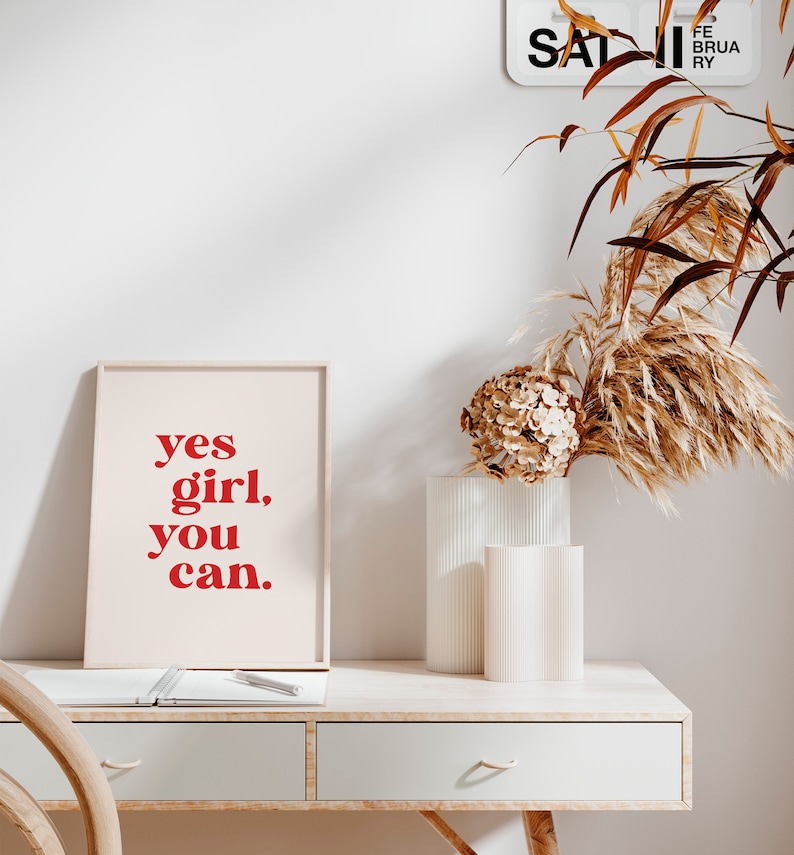 May include: A framed print with the text "yes girl, you can." in red letters on a white background. The print is sitting on a wooden desk with a white notebook and a pen.