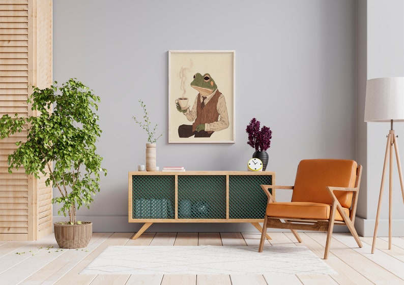 May include: A green frog wearing a brown vest and a tie, holding a cup of coffee, is depicted in a framed illustration. The illustration is hanging on a light gray wall above a wooden cabinet with a green mesh front. A brown leather armchair and a floor lamp are also visible in the image.