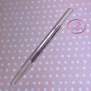 May include: A clear plastic cake decorating tool with a double-ended design. The tool is approximately 12 inches long and has a pointed tip on one end and a rounded tip on the other end.