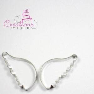 May include: Two silver metal angel wing cookie cutters. The wings have a scalloped edge.
