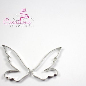 May include: Two silver metal cookie cutters shaped like angel wings. The wings are detailed with a feathered design.