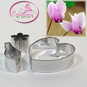 May include: Three silver-colored metal cookie cutters in the shapes of a leaf, a flower, and a heart. The image also includes a pink and purple flower and the text "Creations by Edith" in a pink oval.