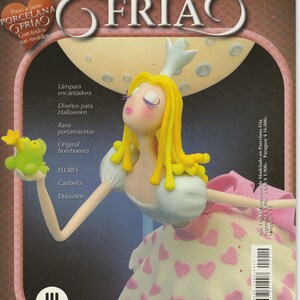 May include: A magazine cover featuring a porcelain doll with blonde hair and a pink dress. The doll is holding a frog and a flower. The magazine cover is titled "Proyectos de modelado en porcelana fria" which translates to "Projects of modeling in cold porcelain".