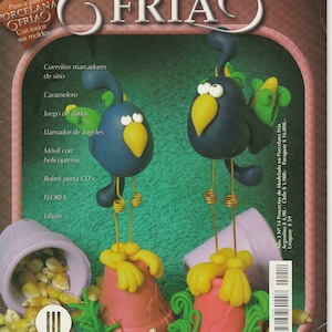 May include: A book cover with the title "Proyectos de modelado en Porcelana Fría" in a decorative font. The cover features two blue and yellow birds with green accents, standing on pink and yellow bases. The birds are made of cold porcelain and are surrounded by yellow seeds.