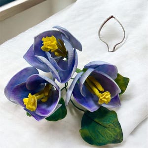 May include: A bouquet of four purple and blue sugar flowers with yellow centers and green leaves. The flowers are arranged in a cluster and are sitting on a white surface.
