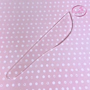 May include: A clear plastic cake decorating tool shaped like a knife with a rounded tip. The tool is on a pink background with white polka dots.