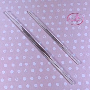 May include: Two clear acrylic cake decorating tools, one longer than the other, on a pink background with white polka dots.