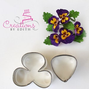 May include: Two metal cookie cutters for making fondant flowers. One cutter is shaped like a three-leaf clover and the other is shaped like a heart. The cutters are on a white surface.