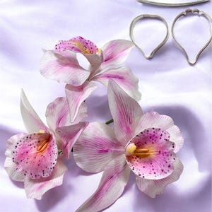 Może przedstawiać: Trzy różowe i białe kwiaty orchidei z fioletowymi plamkami. Kwiaty są wykonane z lukru i służą do dekorowania ciast. Na zdjęciu widać również trzy metalowe foremki do ciasteczek do robienia kwiatów orchidei.