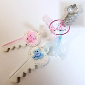 May include: Two white key-shaped cake toppers with pink and blue teddy bears on them. The key toppers are tied with pink and blue ribbons. The text "Creations by Edith" is visible on a white tag.