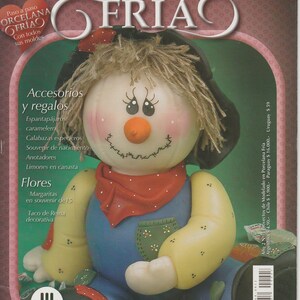 May include: A magazine cover featuring a handmade scarecrow figurine made of cold porcelain. The scarecrow is wearing a red bandana, blue overalls, and a yellow straw hat. The magazine cover is titled "Proyectos de modelado en Porcelana Fría" which translates to "Cold Porcelain Modeling Projects".