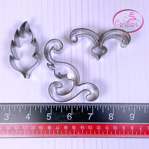 Könnte beinhalten: Drei Metall-Ausstecher, zwei sind verzierte Wirbel und einer ist eine Blattform. Die Ausstecher befinden sich auf einer weißen Oberfläche mit einem Lineal als Maßstab.
