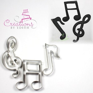 May include: Set of four metal cookie cutters shaped like musical notes. The cutters include a treble clef, a whole note, a half note, and a quarter note. The cutters are silver in color and are arranged on a white surface.